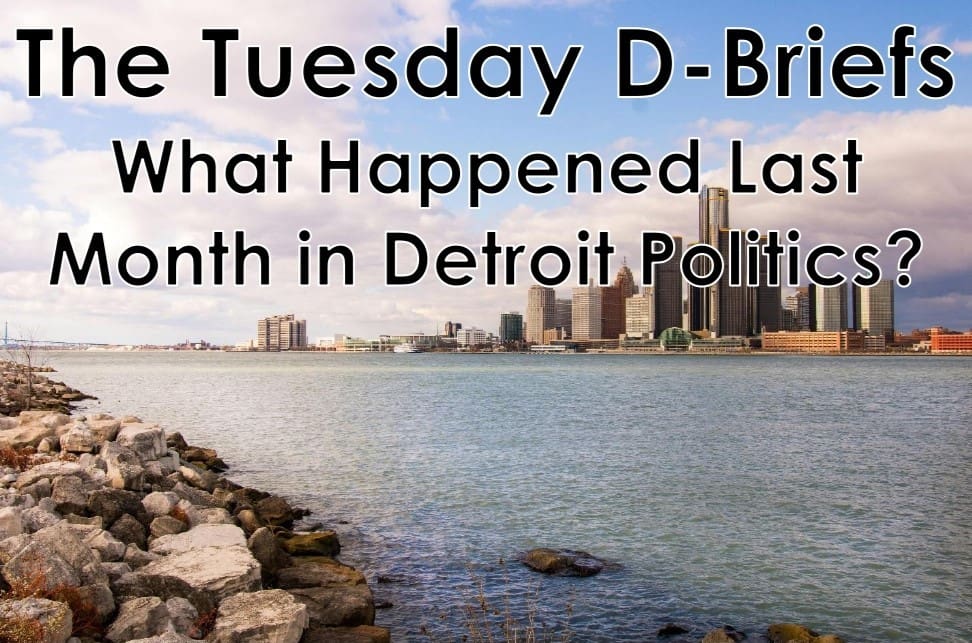 Aerial view of the Detroit skyline across the Detroit River with rocky shoreline in the foreground, overlaid with the text "The Tuesday D-Briefs: What Happened Last Month in Detroit Politics?"