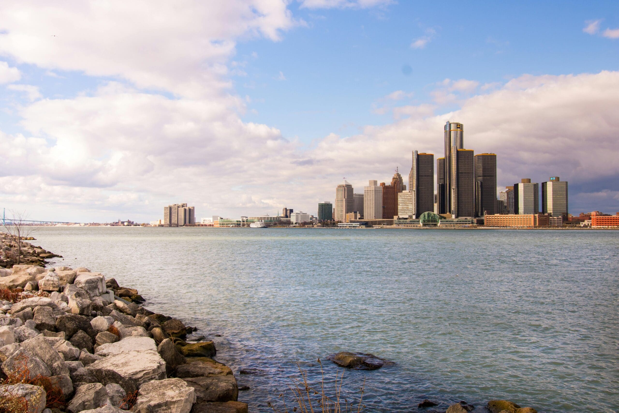 View of the Detroit skyline across the Detroit River, with the Renaissance Center towers visible on the right and the Ambassador Bridge in the far left background, under a partly cloudy sky.