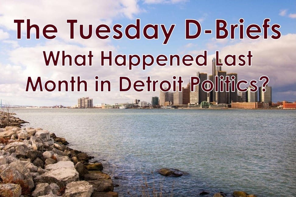 Aerial view of the Detroit skyline across the Detroit River with rocky shoreline in the foreground, overlaid with red text "The Tuesday D-Briefs: What Happened Last Month in Detroit Politics?"