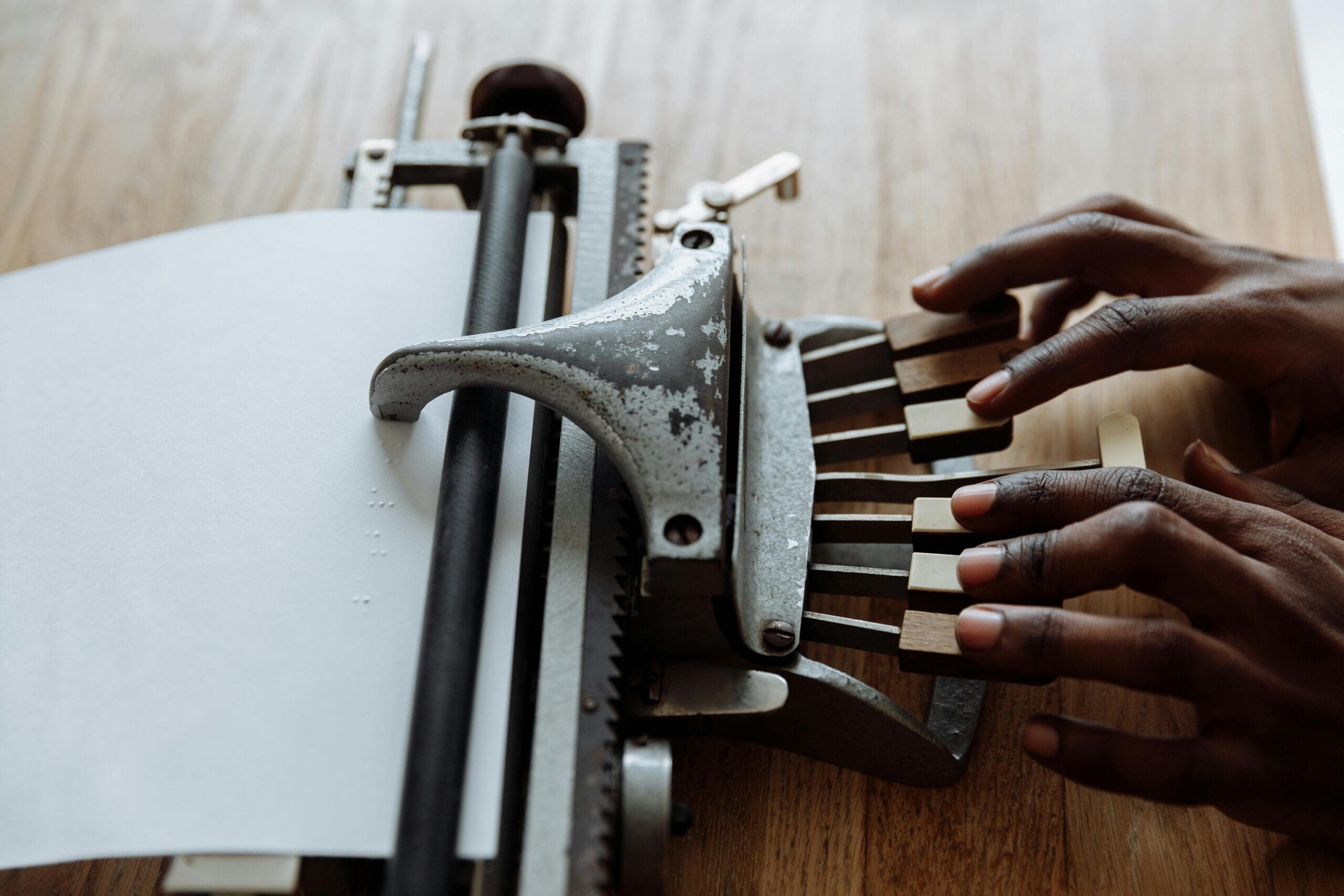 Close-up of hands typing on a metal Braille typewriter, embossing raised dots onto a sheet of paper.